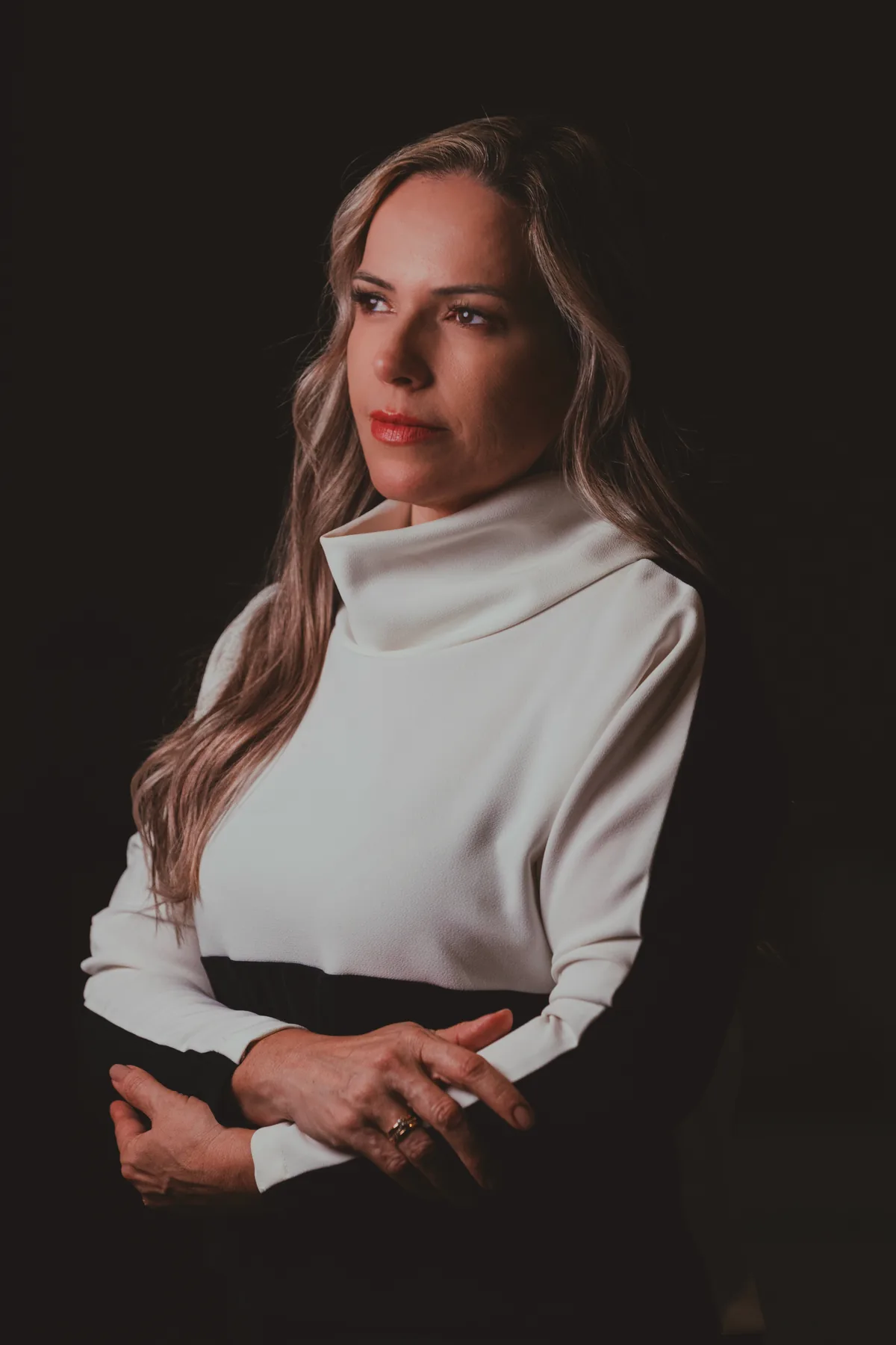 Retrato em meio corpo da Dra. Vanessa Benetti Di Sessa, blusa branca de gola alta, olhar para o lado, braços cruzados, em estúdio escuro