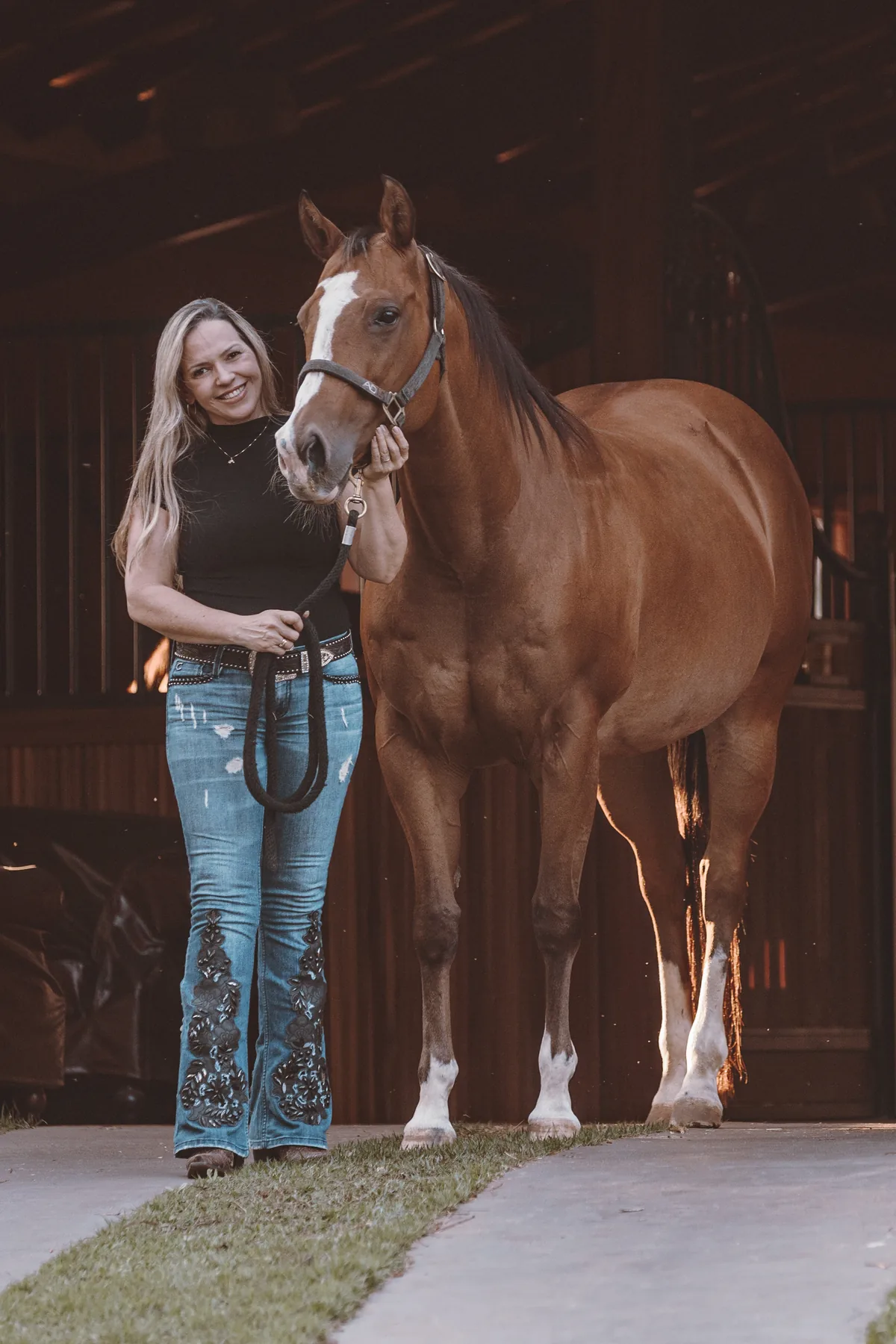 Dra. Vanessa Benetti Di Sessa de corpo inteiro ao lado de um cavalo alazão com cabresto, em uma cocheira iluminada